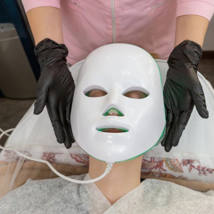 White glossy Light Therapy LED Face Mask held by gloved hands on a person's face with eye, nose, and mouth openings