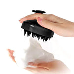 Black silicone Scalp Care Brush with soft triangular tips and ergonomic handle held above hand with white foamy shampoo